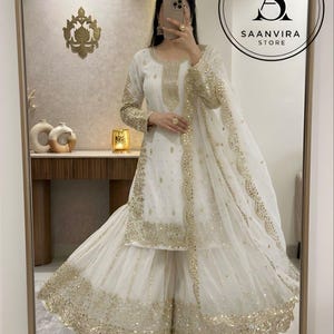 Op de afbeelding: Witte en gouden etnische kleding ensemble. De outfit bevat een tuniek met lange mouwen, een broek met wijde pijpen en een bijpassende sjaal. Gouden pailletten en borduursels sieren de halslijn, mouwen en zoom. Saanvira Store logo.