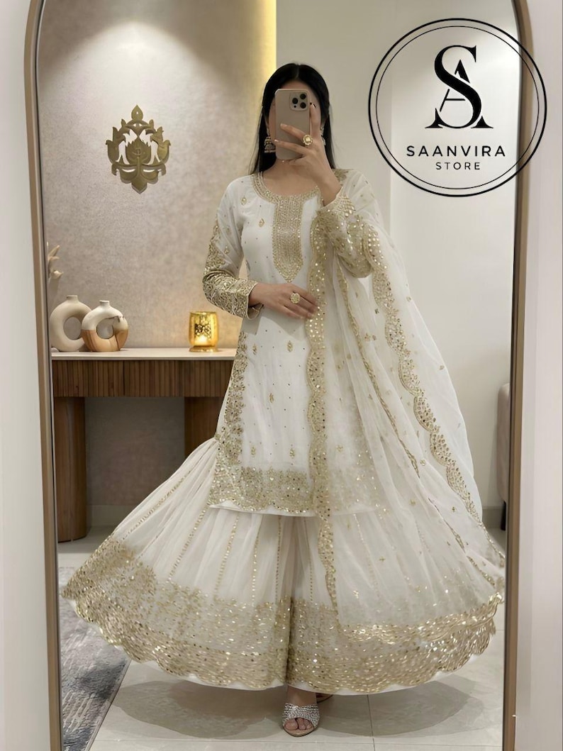 Op de afbeelding: Witte en gouden etnische kleding ensemble. De outfit bevat een tuniek met lange mouwen, een broek met wijde pijpen en een bijpassende sjaal. Gouden pailletten en borduursels sieren de halslijn, mouwen en zoom. Saanvira Store logo.