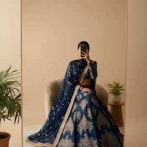 May include: A navy blue lehenga with gold embroidery, a matching dupatta, and a long-sleeved blouse. The outfit is reflected in a large mirror. The Saanvira Store logo is in the upper right corner. The outfit is suitable for special occasions.