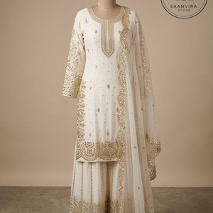 Op de afbeelding: Ivoor kleurige salwar kameez set met ingewikkelde gouden borduursels en versieringen. De set bevat een tuniek met lange mouwen, een broek met wijde pijpen en een transparante dupatta. Het Saanvira Store logo is zichtbaar.