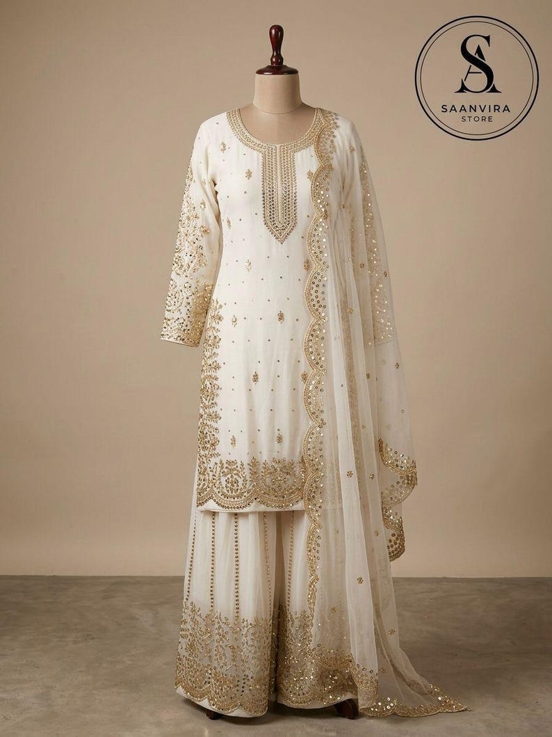 Op de afbeelding: Ivoor kleurige salwar kameez set met ingewikkelde gouden borduursels en versieringen. De set bevat een tuniek met lange mouwen, een broek met wijde pijpen en een transparante dupatta. Het Saanvira Store logo is zichtbaar.