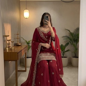 May include: A burgundy salwar kameez set with gold embroidery. The outfit features a long-sleeved top, wide-legged trousers, and a matching dupatta. The image also shows a mirror with the text "SAANVIRA STORE".