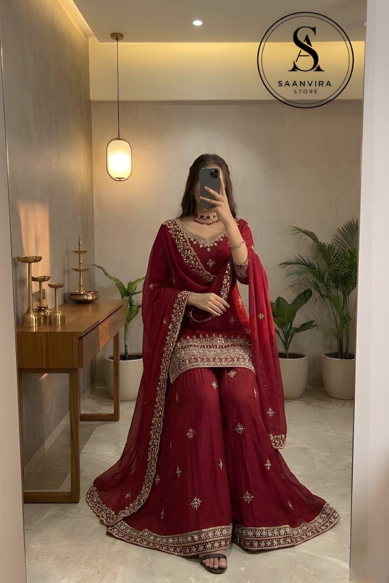 May include: A burgundy salwar kameez set with gold embroidery. The outfit features a long-sleeved top, wide-legged trousers, and a matching dupatta. The image also shows a mirror with the text "SAANVIRA STORE".