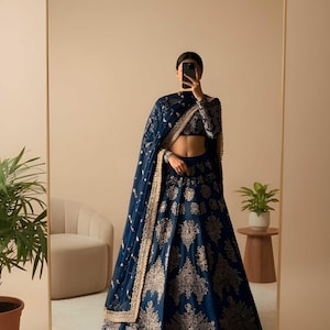 May include: A navy blue lehenga with gold embroidery, long sleeves, and a matching dupatta. The lehenga features intricate floral patterns. The image is reflected in a large mirror. The Saanvira Store logo is visible in the top right corner.