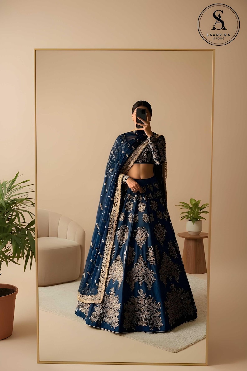 May include: A navy blue lehenga with gold embroidery, long sleeves, and a matching dupatta. The lehenga features intricate floral patterns. The image is reflected in a large mirror. The Saanvira Store logo is visible in the top right corner.