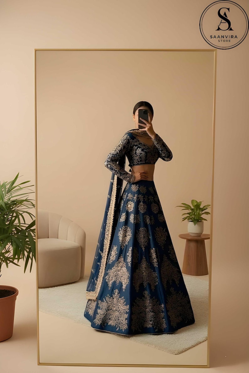 May include: A navy blue lehenga with gold floral embroidery. The long-sleeved blouse and skirt feature intricate patterns. The outfit is reflected in a large mirror, with the Saanvira Store logo visible in the top right corner.