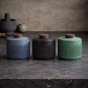Op de afbeelding: Drie keramische potten in blauw, zwart en groen, elk met een geribbeld ontwerp en een houten deksel. De potten zijn cilindrisch en staan op een donkere ondergrond.