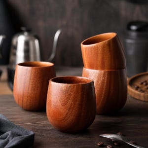 Op de afbeelding: Vier houten bekers met een natuurlijke houtnerf afwerking zijn gerangschikt op een donker houten oppervlak. De bekers hebben een afgerond, stamlloos ontwerp, met een gladde, gepolijste buitenkant. De bekers zijn gestapeld en geplaatst in de buurt van koffiebonen en een lepel.