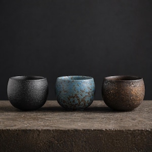 May include: Three handcrafted ceramic bowls in a row. The bowls are in a dark color palette, with a black, blue, and brown bowl. The bowls have a textured surface and a rounded shape. The bowls are likely for serving food or as decorative items.