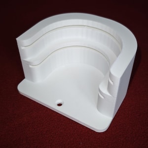 May include: A white, curved bracket with a flat base and a hole for mounting. The bracket has a layered design with three recessed sections. The object is made of a solid, opaque material and sits on a red surface.