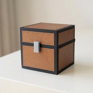 May include: A square, brown chest with a black border and a gray clasp. The chest has a textured appearance and is sitting on a white surface. The chest is a decorative item.