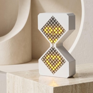 May include: A white, hourglass-shaped digital clock featuring two diamond-shaped LED display panels. Each panel shows a glowing yellow pixelated heart design. The device has a smooth, matte finish and a geometric, angular frame structure.