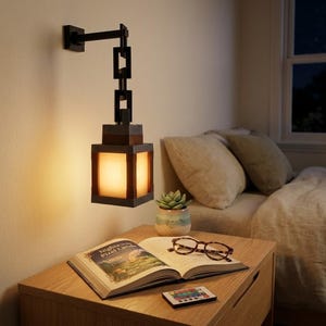 May include: A dark brown, geometric wall-mounted lamp with a rectangular shade, illuminating a warm glow. The lamp hangs above a wooden nightstand with an open book, glasses, a remote, and a small potted succulent.