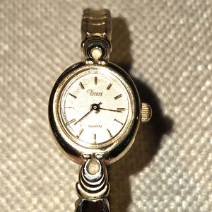 May include: A gold-tone Timex quartz wristwatch featuring a round case with a matching gold-tone dial. The watch has a metallic link bracelet with a decorative design connecting to the case. The face displays the Timex logo and the word QUARTZ.