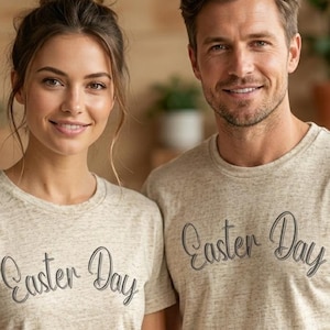 May include: Two people wearing matching beige t-shirts with the text "Easter Day" in a script font. The shirts have a heathered texture. The background includes plants in pots and a wooden wall.