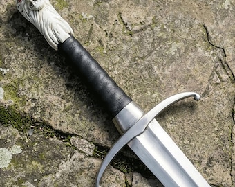 May include: A sword featuring a silver-colored metal blade and a crossguard. The handle is wrapped in black textured material and topped with an off-white pommel sculpted into the shape of a roaring lion head with small red-colored eyes.