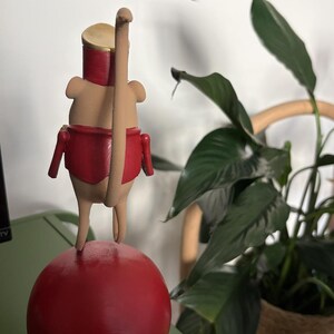May include: A tan-colored mouse figurine wearing a red jacket and a gold-colored hat, standing on a large red sphere. The mouse has a long, thin tail extending upward and is positioned in a vertical stance on top of the round red base.