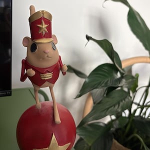May include: A figurine of a light brown mouse wearing a red and gold military-style jacket and a matching hat with a gold star. The mouse is perched on top of a large, smooth red sphere that features a prominent gold star on its front surface.