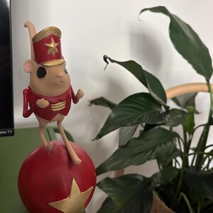 May include: A figurine of a light tan mouse wearing a red and gold military-style jacket and a matching hat with a gold star. The mouse is balanced on top of a large red sphere that features a prominent gold star on its front surface.