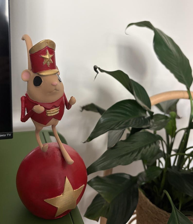 May include: A figurine of a light tan mouse wearing a red and gold military-style jacket and a matching hat with a gold star. The mouse is balanced on top of a large red sphere that features a prominent gold star on its front surface.