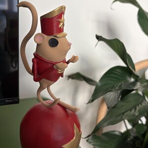 May include: A tan mouse figurine dressed in a red and gold military-style jacket and matching hat with a gold star. The mouse has a black circular eye and is perched on top of a large, round, red ball decorated with a gold star on its side.