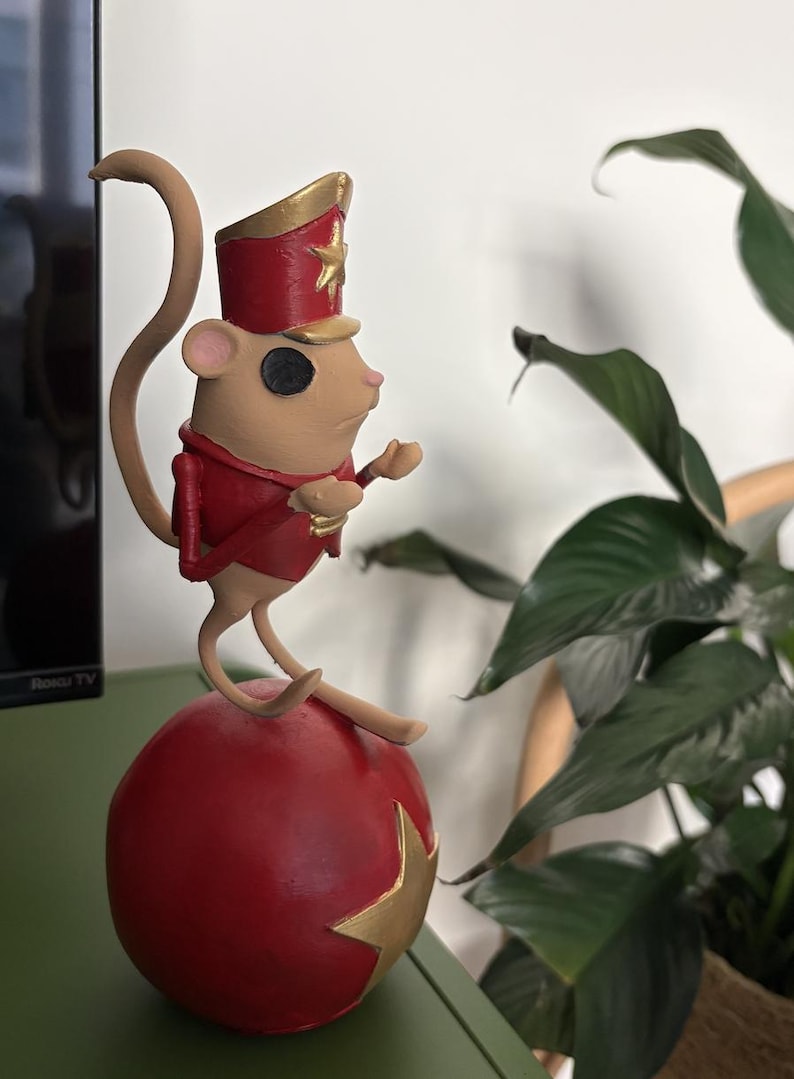 May include: A tan mouse figurine dressed in a red and gold military-style jacket and matching hat with a gold star. The mouse has a black circular eye and is perched on top of a large, round, red ball decorated with a gold star on its side.