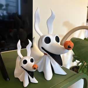May include: Two white ghost dog figurines with long ears, black oval eyes, and wide black mouths. Each figure features a small orange pumpkin nose. The larger dog has a red collar around its neck, while the smaller dog is positioned to its left.
