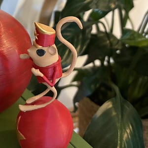 May include: A small beige mouse figurine wearing a red jacket and a matching red hat with gold trim. The mouse is balanced on top of a large, smooth red sphere. The figure has a long, curved tail and is posed in a standing position on the ball.