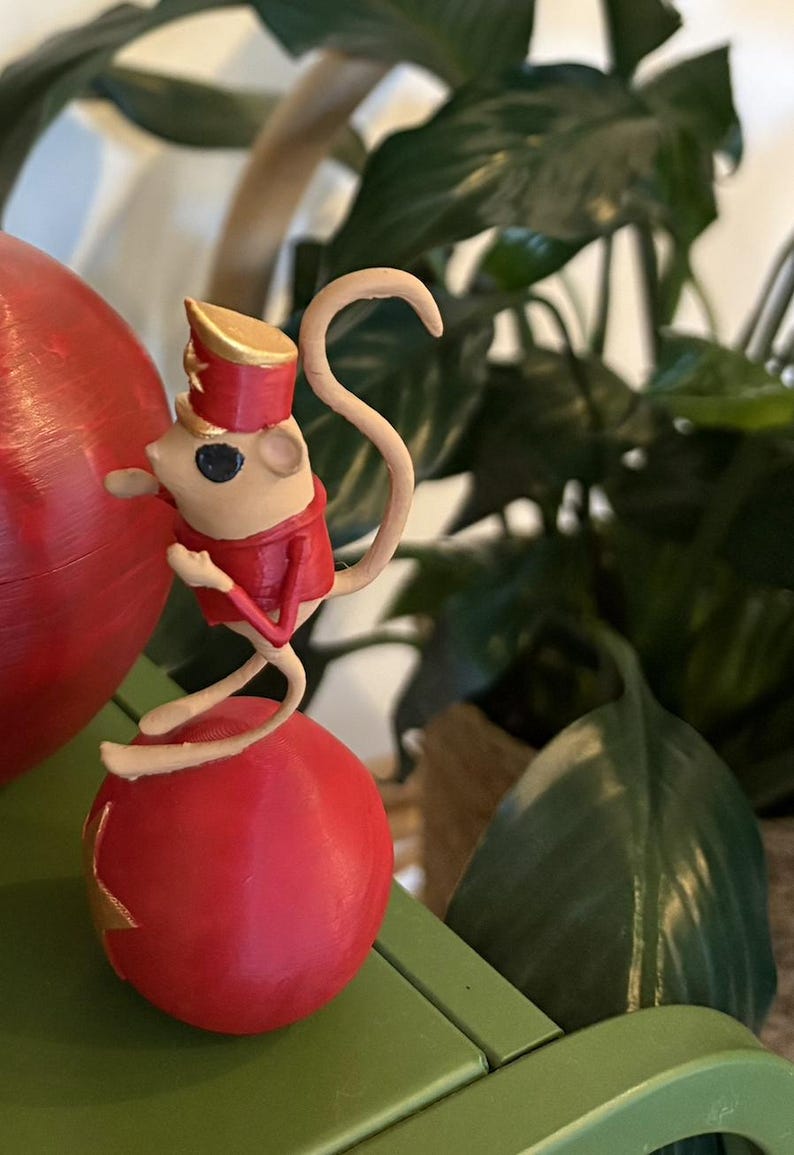 May include: A small beige mouse figurine wearing a red jacket and a matching red hat with gold trim. The mouse is balanced on top of a large, smooth red sphere. The figure has a long, curved tail and is posed in a standing position on the ball.