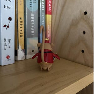 May include: A small tan mouse figurine wearing a red jacket with black trim and a gold-colored hat. The mouse has a long, curved tail and is positioned facing away from the viewer, standing on a light-colored wooden shelf next to several books.