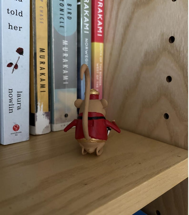May include: A small tan mouse figurine wearing a red jacket with black trim and a gold-colored hat. The mouse has a long, curved tail and is positioned facing away from the viewer, standing on a light-colored wooden shelf next to several books.