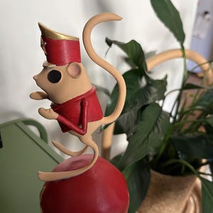 May include: A beige mouse figurine wearing a red uniform jacket and a matching red hat with gold trim. The mouse is balanced on top of a large, smooth red sphere. The figure has a long, curved tail and is posed in a standing position on the ball.