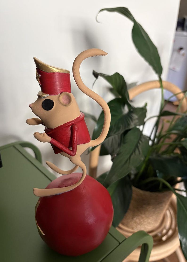 May include: A beige mouse figurine wearing a red uniform jacket and a matching red hat with gold trim. The mouse is balanced on top of a large, smooth red sphere. The figure has a long, curved tail and is posed in a standing position on the ball.