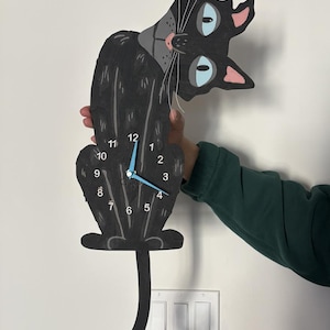 May include: A black cat-shaped wall clock with a whimsical design. The clock features a gray collar, blue eyes, and pink inner ears. The clock hands are light blue. The clock is held against a white wall.