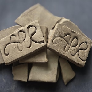 May include: A collection of rectangular, light brown clay tiles with a matte, earthy texture. Two of the tiles feature a prominent, hand-carved, continuous swirling line design that forms an abstract, symmetrical pattern across the surface.