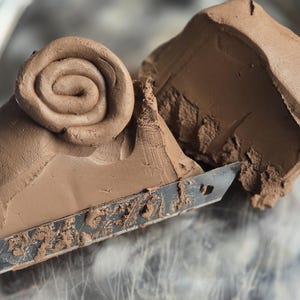 May include: Close-up of brown clay with a spiral design, cut with a utility knife. The clay is a rich, earthy tone, and the knife blade is silver. The image highlights the texture and form of the clay.