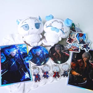 May include: A collection of anime-themed merchandise, including two white plush toys with blue accents, stickers, buttons, keychains, and a holographic print. The items feature character illustrations in various poses and styles.