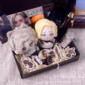 May include: A gift box containing two plush dolls with blonde and grey hair, several character stickers, a circular pin, and an acrylic standee featuring a blonde character. The items are nestled in shredded paper filler within a black cardboard box.