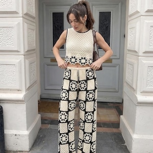 以下が含まれることがあります： クリーム色のクロシェ編みトップスとパンツのセット。トップスはノースリーブで、黒いトリムと裾に花柄があります。パンツは、黒い花のモチーフの市松模様のデザインが特徴です。この衣装は、スタイリッシュな手作りのアンサンブルです。