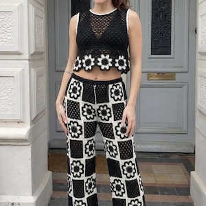 May include: Black and white crochet two-piece set. The top is a sleeveless crop top with a white trim and a floral design. The pants are wide-legged with a checkerboard pattern of black and white floral squares. The outfit is handmade.