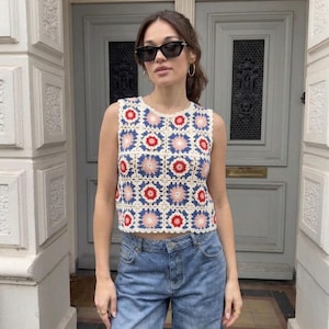 Peut inclure: Un haut sans manches en crochet avec un motif floral rouge, bleu et blanc. Il est associé à un jean en denim bleu clair. La tenue est portée devant une porte grise claire.