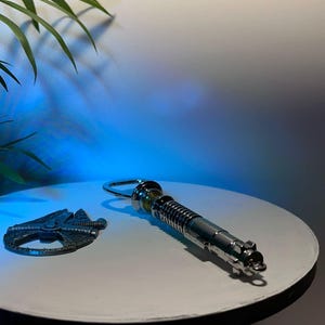 Puede incluir: Un abrebotellas de sable de luz plateado y un abrebotellas del Halcón Milenario sobre una superficie blanca. El abridor de sable de luz tiene un anillo para colgar. El abridor del Halcón Milenario es de metal oscuro.