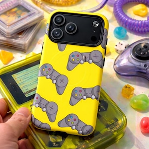May include: A yellow phone case with a pattern of gray video game controllers. The phone case is on top of a yellow handheld gaming device. Other colorful objects are in the background.