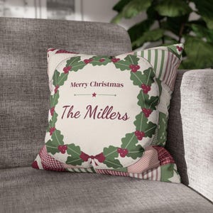 Op de afbeelding: Een vierkant sierkussen met een crèmekleurig middenpaneel voorzien van een groene hulstkrans, rode bessen en de tekst Merry Christmas en The Millers in donkerrood schrift. De rand bestaat uit een patchworkpatroon van rode en groene stoffen.