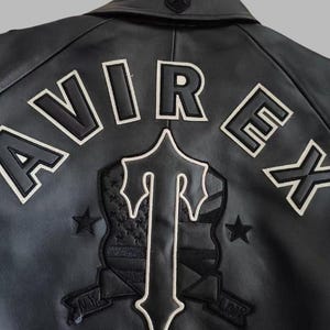 Black Avirex leather jacket featuring bold embroidered Avirex LDN lettering on the back, classic bomber style, ribbed cuffs and hem, premium craftsmanship.