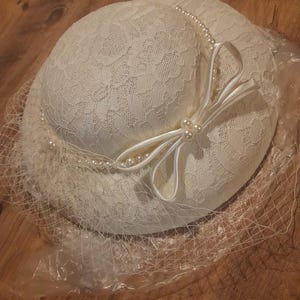 May include: Ivory-colored hat with a delicate lace overlay, adorned with a satin bow and faux pearl accents. A fine mesh veil drapes over the brim. The hat is a classic accessory.