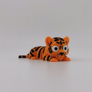 May include: A small, orange tiger figurine in a lying position. The tiger features bold black stripes across its body, head, and tail. It has large, round white eyes with black pupils, a small black nose, and detailed whiskers on its muzzle.