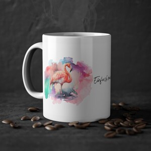 Könnte beinhalten: Weiße Keramik-Tasse mit einem Aquarell-Flamingo-Design in Rosa, Orange und Lila. Die Tasse hat einen schwarzen Henkel und den Text "Einfach mal" in Schreibschrift. Kaffeebohnen sind um die Basis der Tasse verstreut.