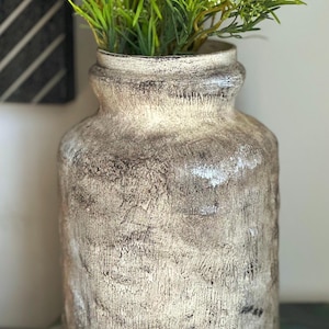 May include: A cylindrical ceramic vase with a textured, weathered finish in shades of cream, beige, and dark brown. The vase features a wide, slightly flared rim and a thick, uneven surface texture. It is filled with green artificial foliage.