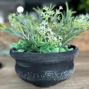 May include: A black, textured ceramic pot filled with greenery and small white flowers. The pot has a decorative band around the middle and sits on a wooden surface. The arrangement includes various shades of green.